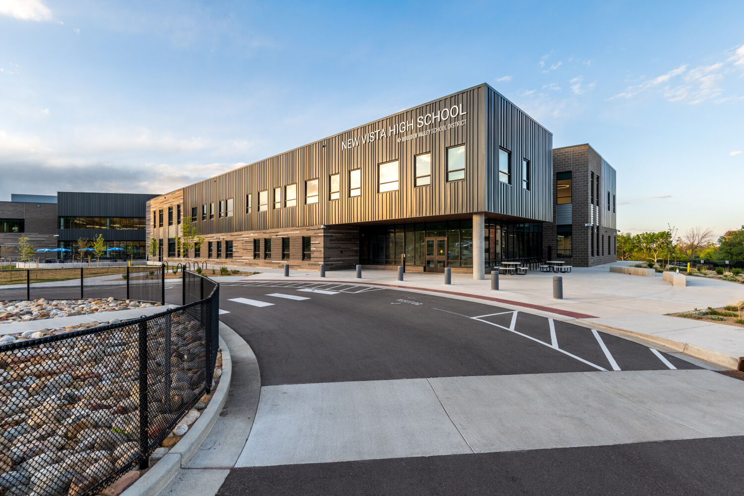 Main entrance of New Vista High School with metal cladding, brick base, recessed entry canopy, large glazing, and a curved drop‑off drive with site landscaping.