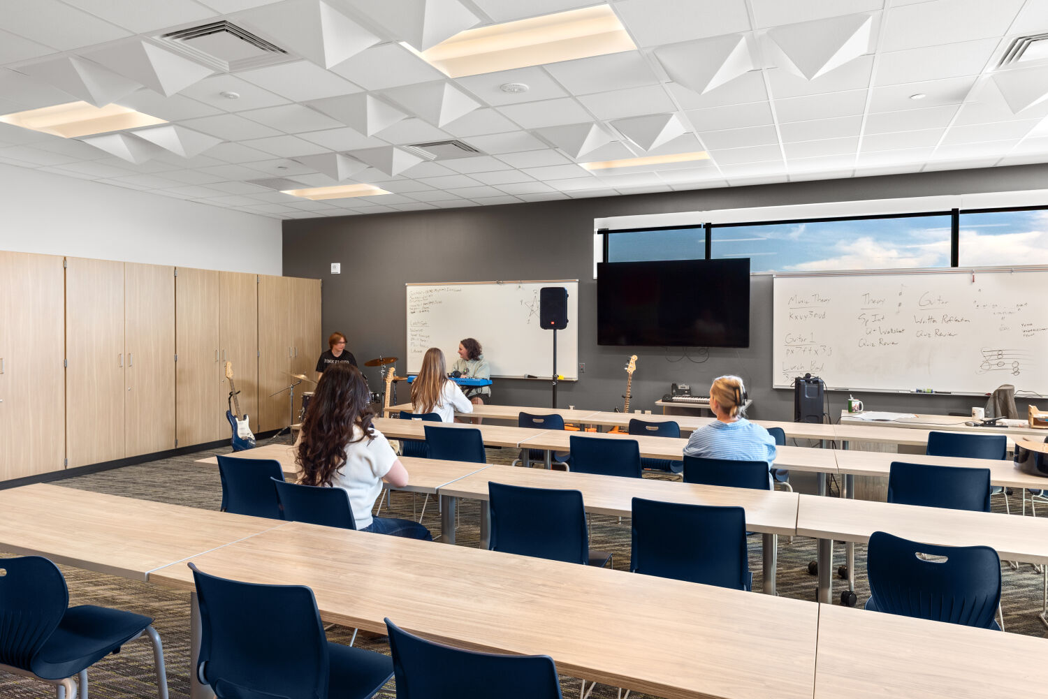 Music classroom with long tables and chairs arranged in rows, whiteboards, acoustic ceiling panels, built‑in storage cabinets, and guitars positioned at the front of the room.