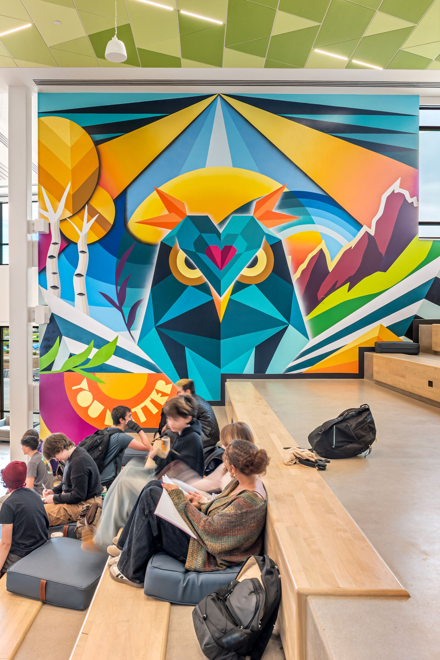 Learning stair with tiered wood seating and integrated cushions, set beside a large, colorful geometric mural depicting an owl with stylized mountains, trees, and sunburst elements, under a ceiling with green acoustic panels.