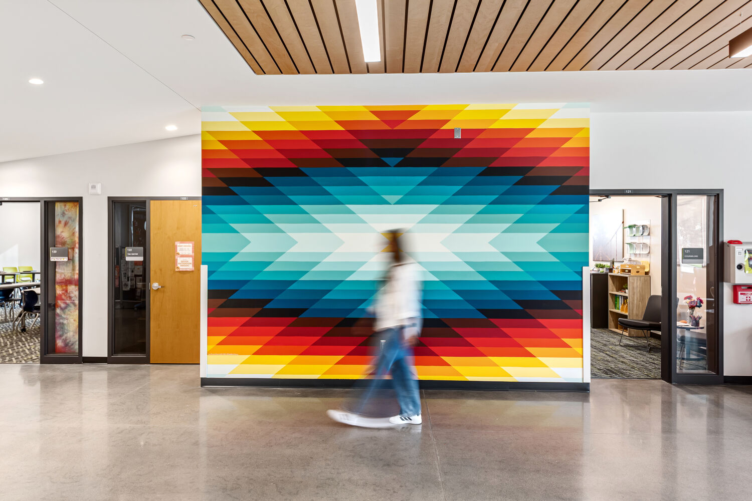 School corridor with a large geometric wall mural in red, yellow, teal, and black tones, polished concrete flooring, wood‑slat ceiling accents, and classroom entrances on both sides.
