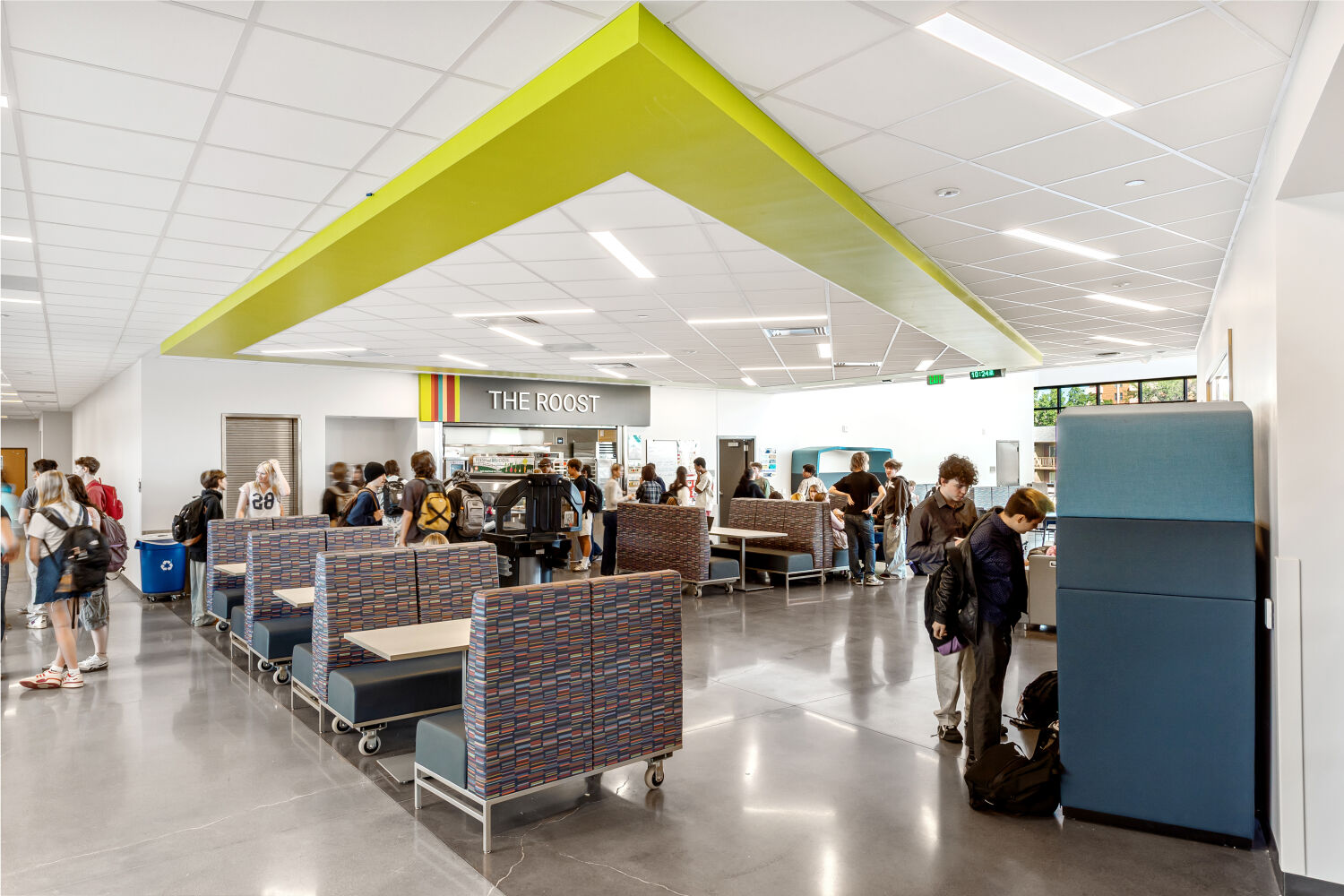 Dining area with patterned booth seating, movable tables, polished concrete flooring, a ceiling feature with bright green angled panels, and a food‑service counter labeled ‘The Roost’ beneath colorful vertical stripes.
