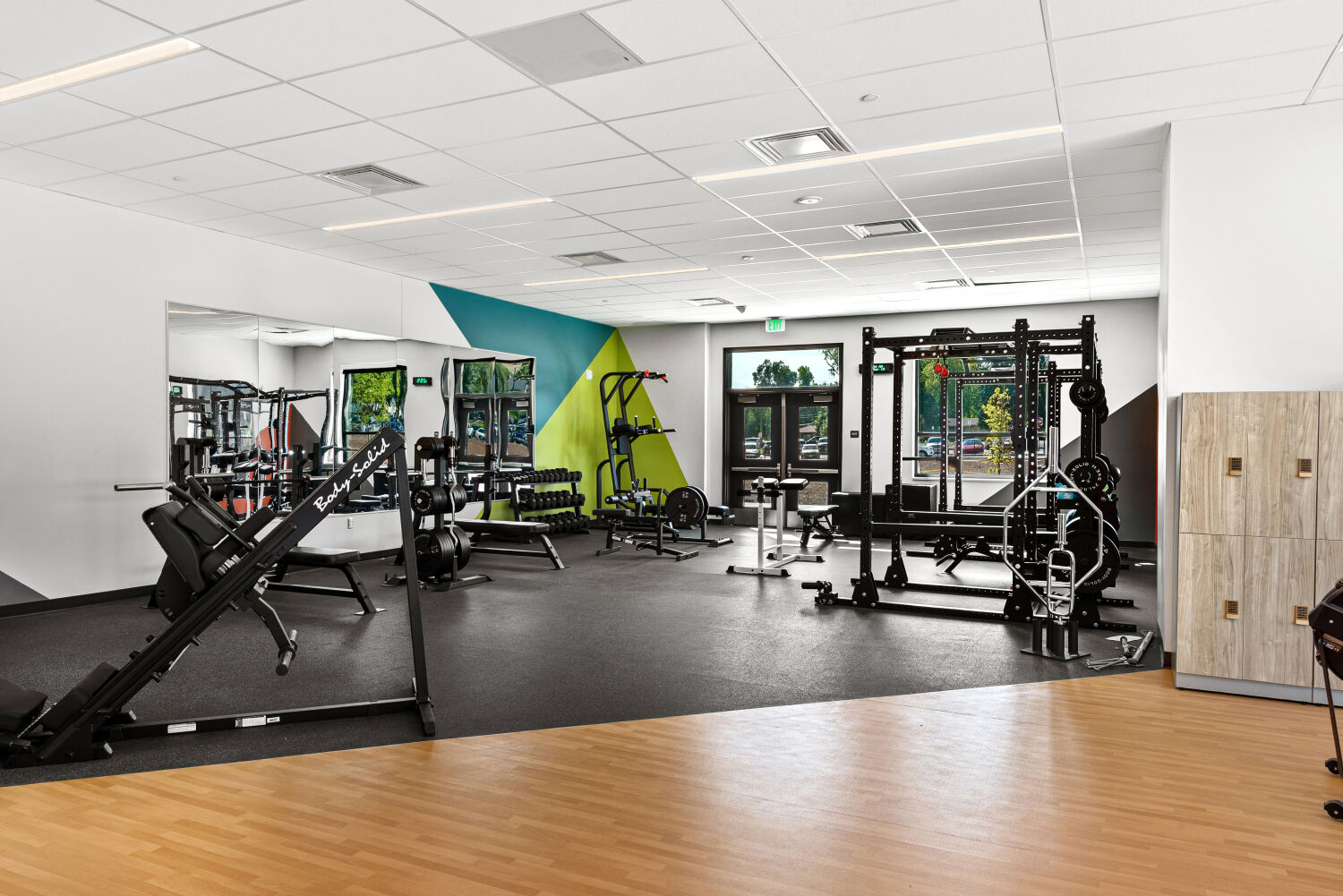 Fitness room with weight‑training equipment including squat racks, dumbbells, benches, and resistance machines, featuring colorful geometric wall accents, large mirrors.