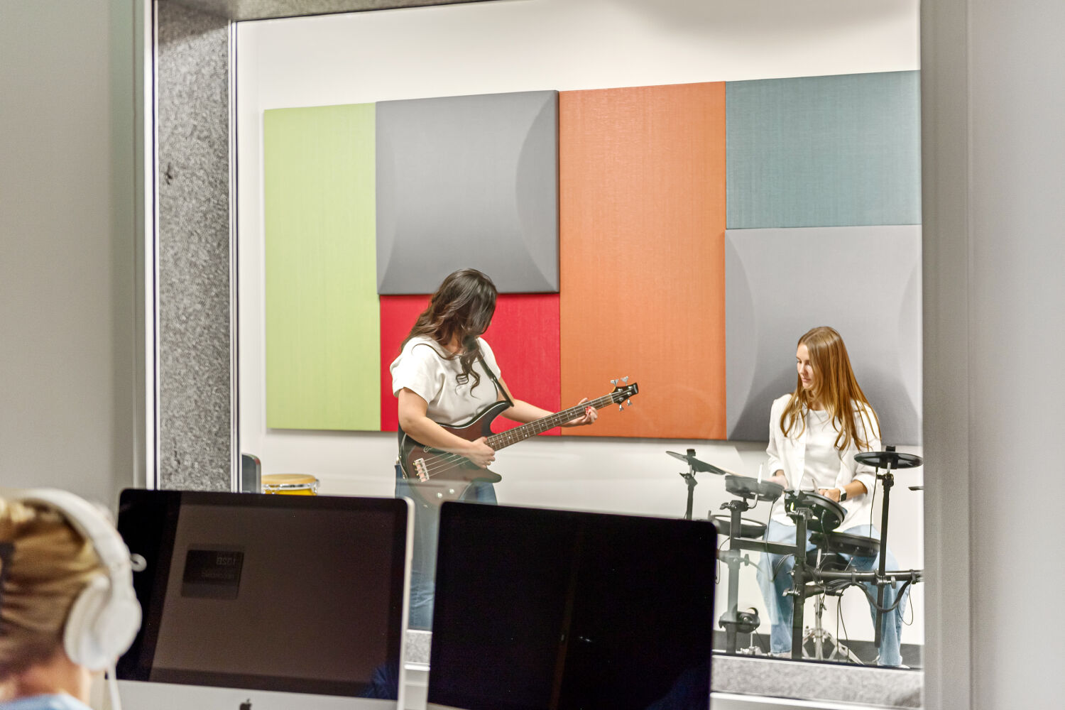 Sound booth with colorful acoustic wall panels, an electric guitar and electronic drum kit positioned against the treated wall, and a window looking into an adjacent workstation area with desktop computers.