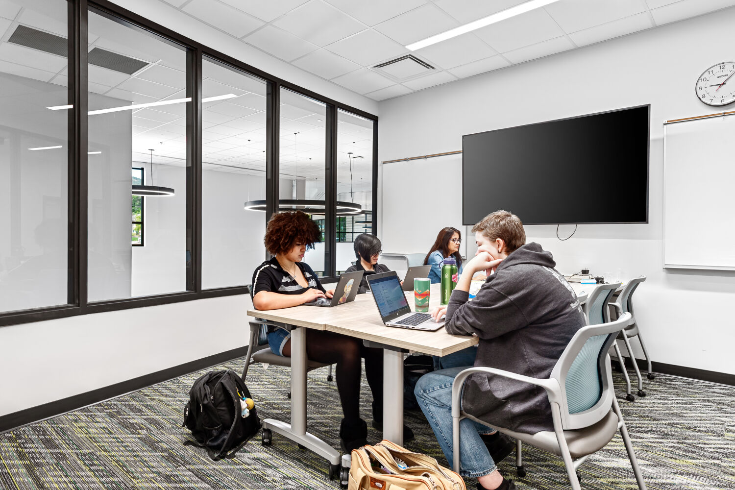 Small‑group classroom with glass interior windows, a wall‑mounted display screen, whiteboards, rectangular table with task chairs, patterned carpet tile flooring, and a ceiling with recessed lighting.