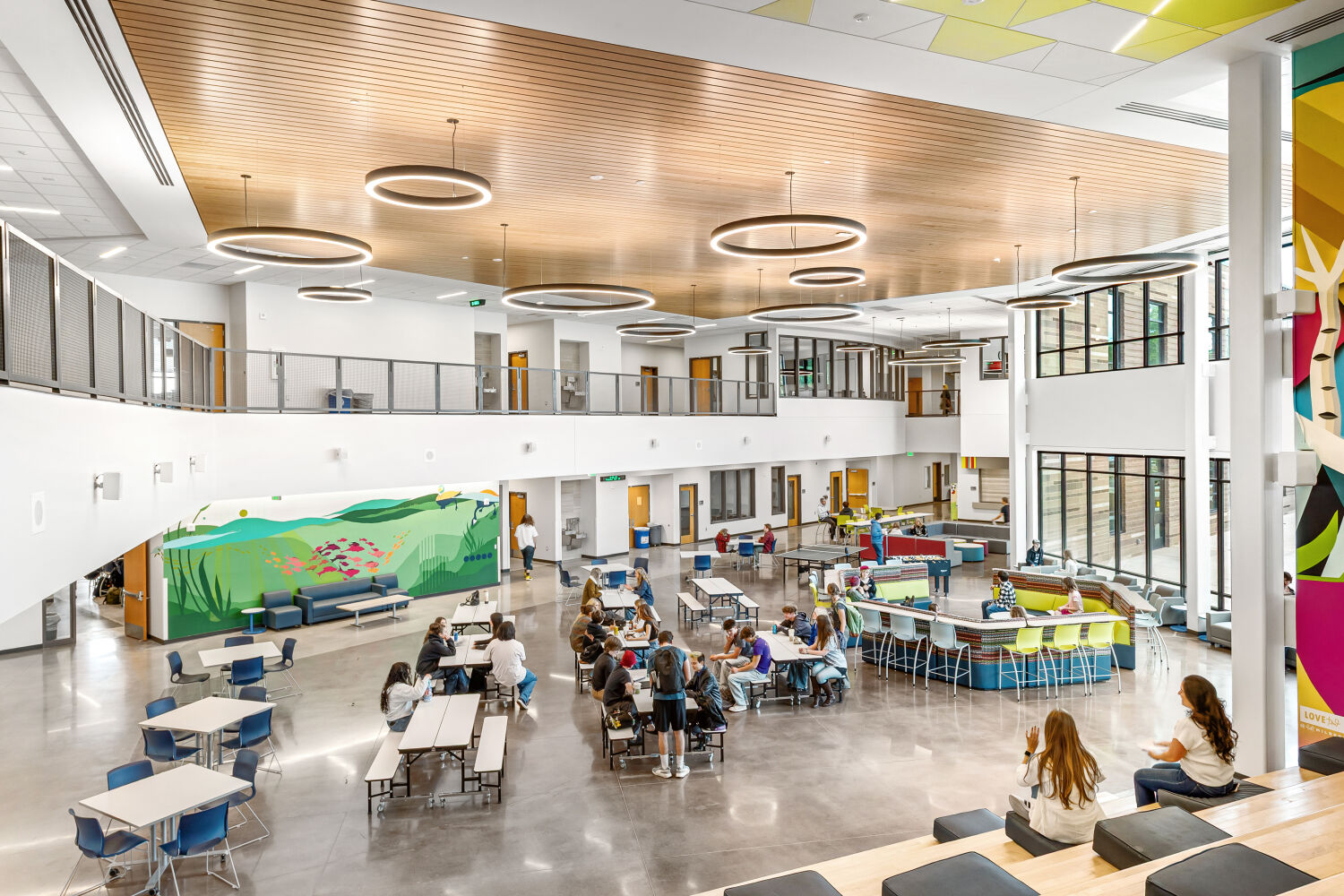 Two‑story school commons with polished concrete flooring, large windows, circular pendant lights, wood‑slat ceiling accents, cafeteria tables, curved lounge seating, and a colorful nature‑themed mural on one wall.