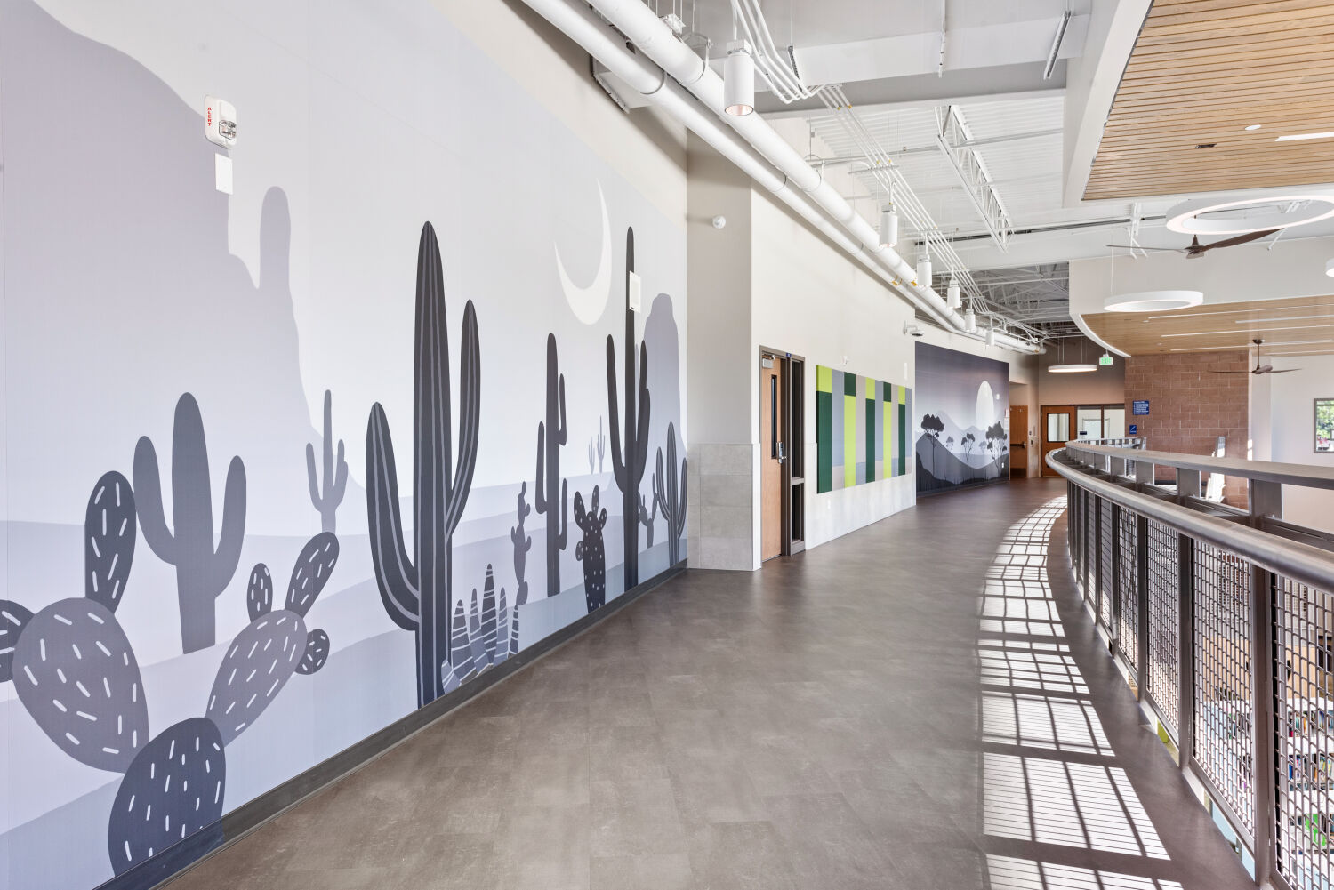 Upper-level corridor with a greyscale desert-themed wall mural featuring cacti and mountains, metal railing overlooking the media center below.
