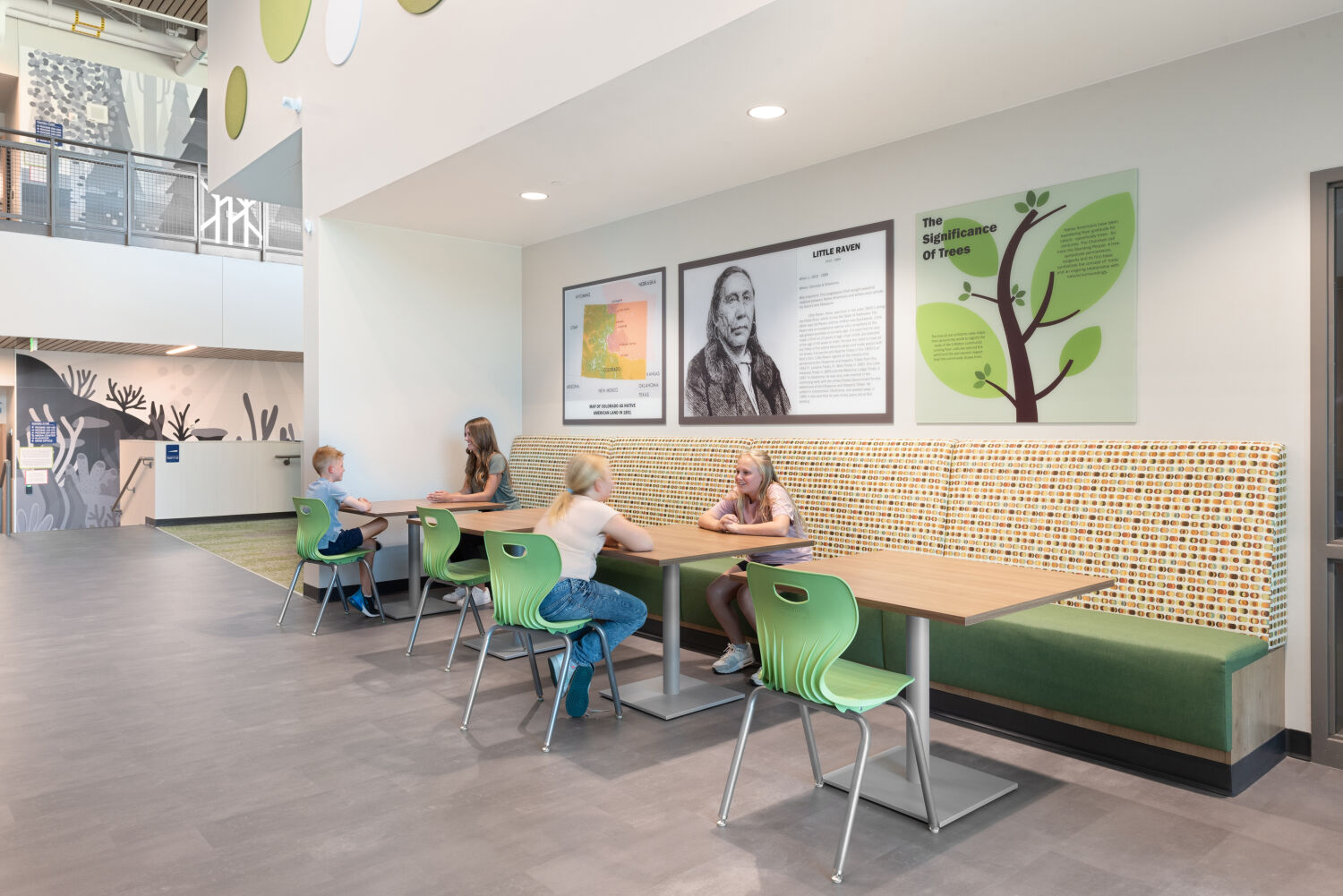 Commons seating area with patterned banquette bench, rectangular tables with green chairs, wall graphics featuring educational panels and a tree illustration.