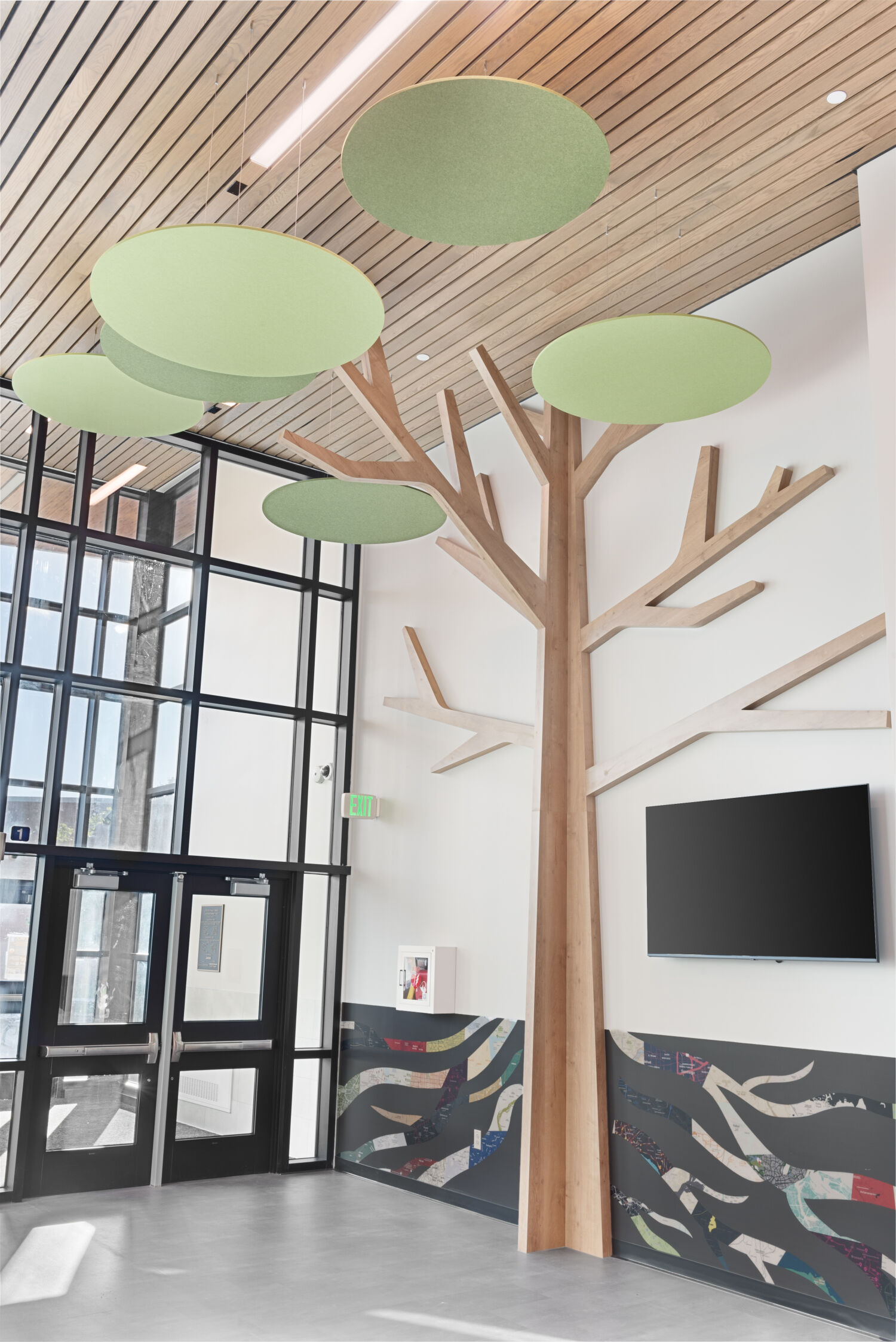 Entry lobby with a tall wood installation shaped like a stylized tree, featuring large circular green acoustic panels overhead, wood‑slat ceiling accents, floor‑to‑ceiling windows, abstract patterned wall panels, and a wall‑mounted display screen.