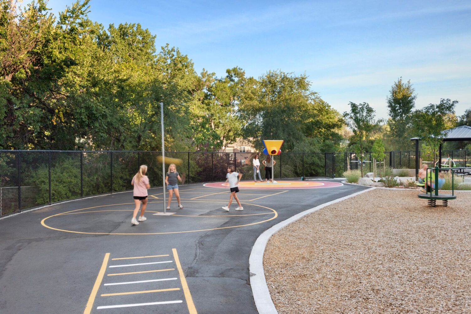 Outdoor play area with an asphalt game court marked with circular play zones and ladder-style lines, a freestanding multi‑hoop basketball feature, perimeter fencing, surrounding trees, and adjacent playground equipment on a wood‑mulch surface.