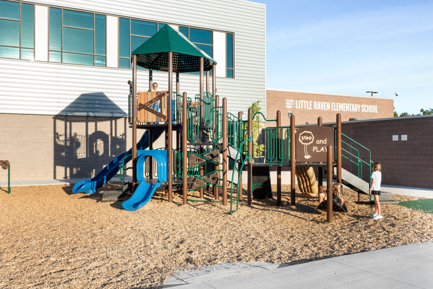 Playground with a multi‑level climbing structure featuring green and brown support posts, blue slides, rope elements, and elevated platforms, set on a wood‑mulch surface beside the exterior of Little Raven Elementary School.