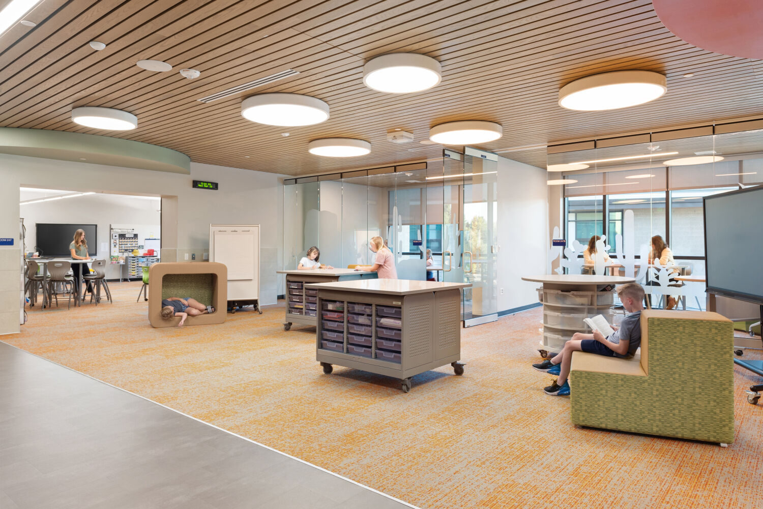 Collaborative learning area with rolling worktables, built‑in storage islands, soft seating nooks, patterned carpet flooring, wood‑slat ceiling with circular light fixtures, and adjacent glass‑walled breakout rooms.
