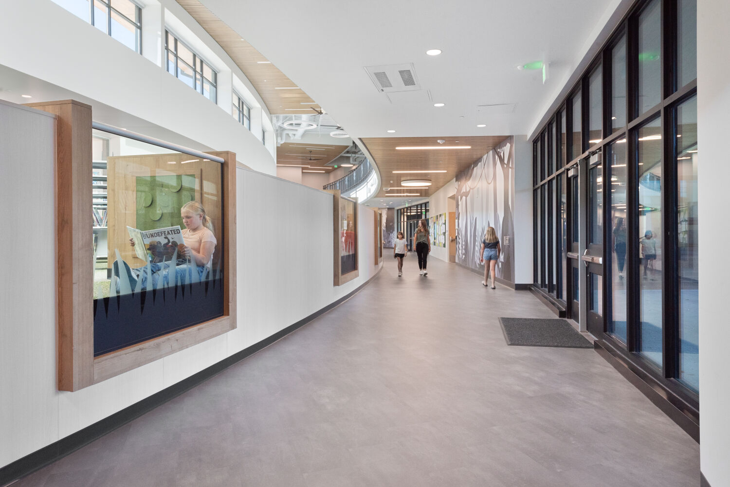 Curved interior corridor with floor‑to‑ceiling glass along one side, framed display windows set into a white wall on the opposite side, wood‑slat ceiling accents with circular light fixtures, and access points to adjacent classrooms and learning areas.
