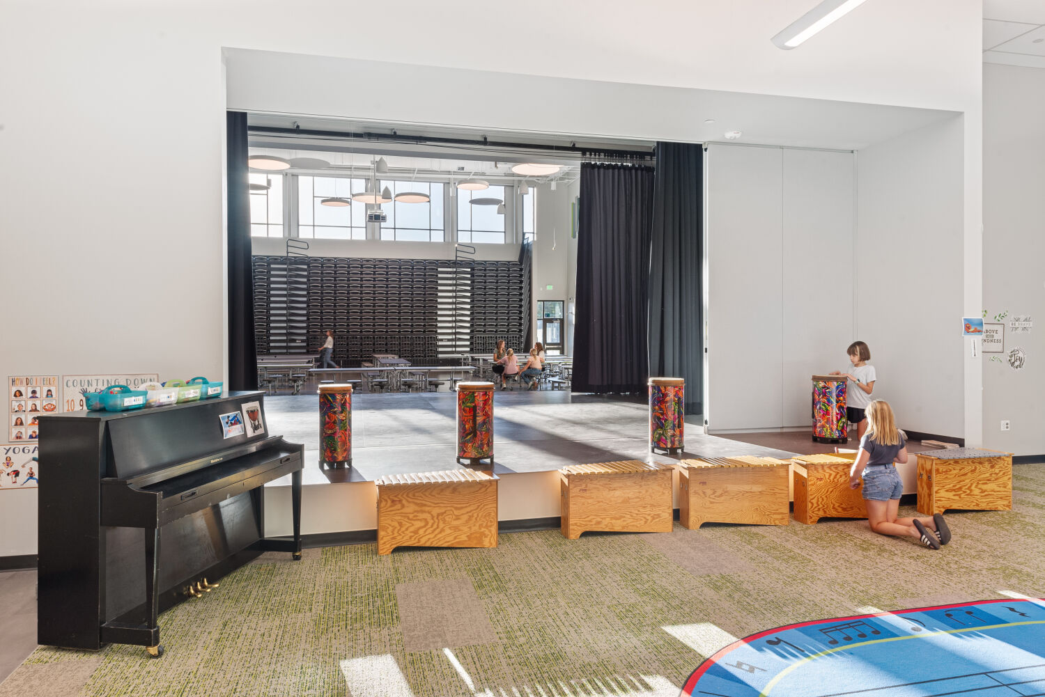 Small performance stage with wood step platforms, colorful hand‑drum instruments arranged along the front edge, black stage curtains, overhead projector, adjacent upright piano, and surrounding carpeted learning area.