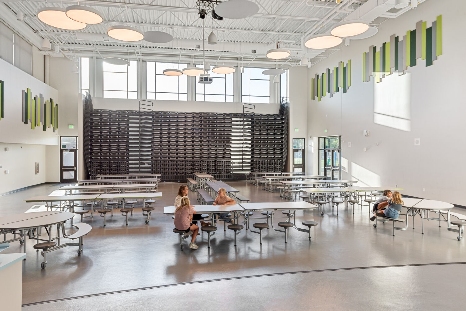 Two‑story cafeteria with large windows, green and gray vertical acoustic panels on the upper walls, retractable bleacher seating along the back wall, movable cafeteria tables with round stools, and polished flooring.