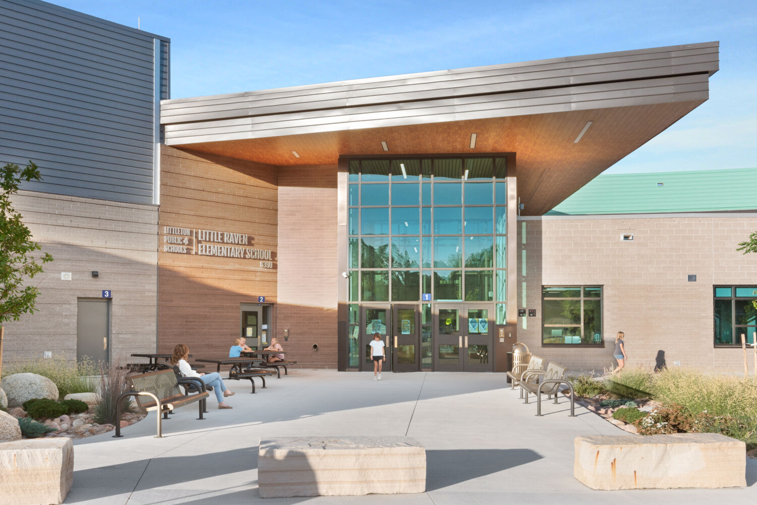 Main entrance of Little Raven Elementary School featuring a tall glass façade, angled roof canopy with wood soffit, landscaped seating areas with concrete benches, and paved walkways leading to the front doors.
