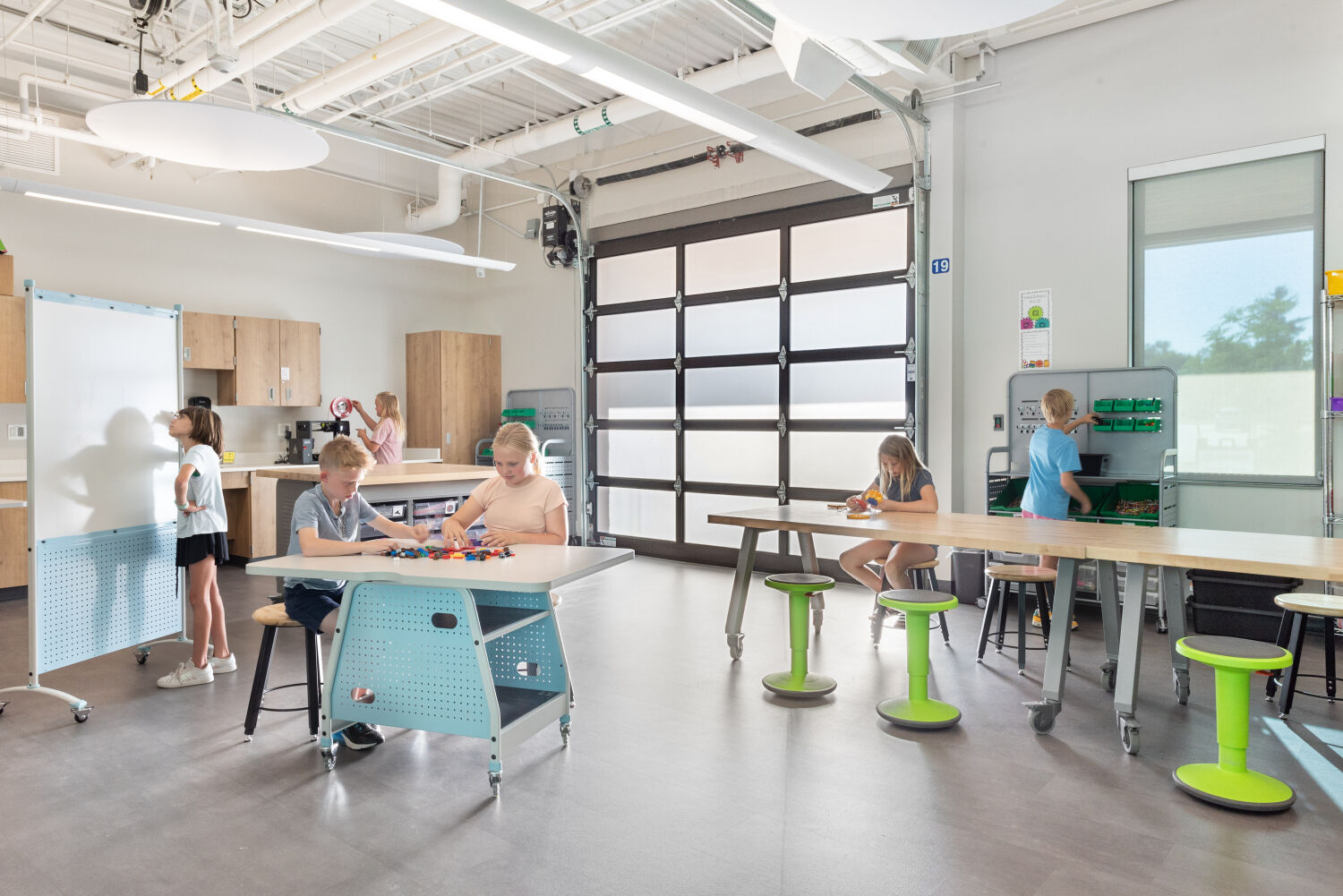 Maker space with high worktables and stools, mobile whiteboard, wood cabinetry with storage, garage‑style glass door, supply shelves, exposed ceiling with suspended lighting, and a combination of hard‑surface and carpet flooring.