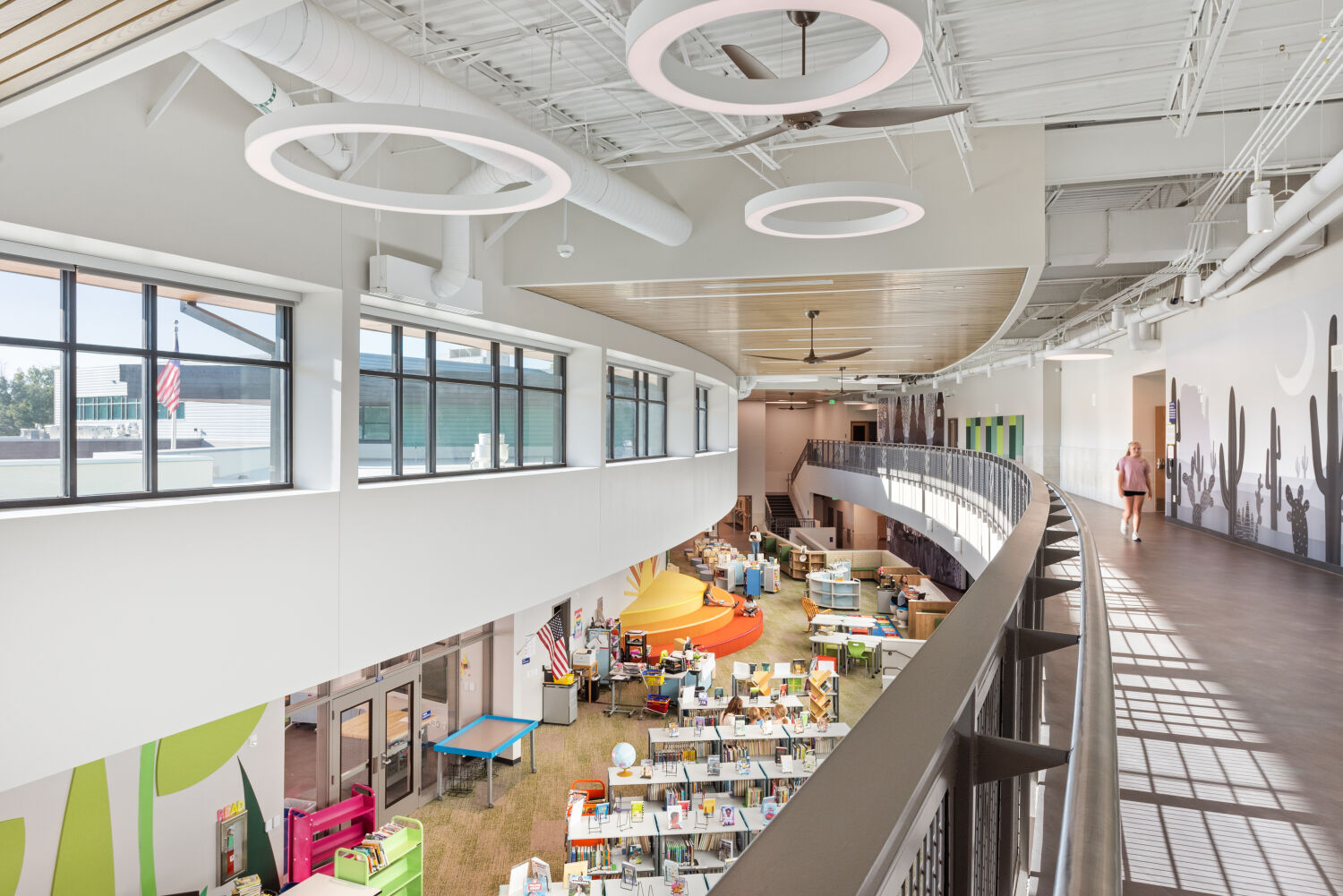 Upper‑level walkway overlooking the media center, featuring a curved metal railing, wood‑slat ceiling accents with large circular light fixtures, floor‑to‑ceiling windows along one wall, and the open media center below.