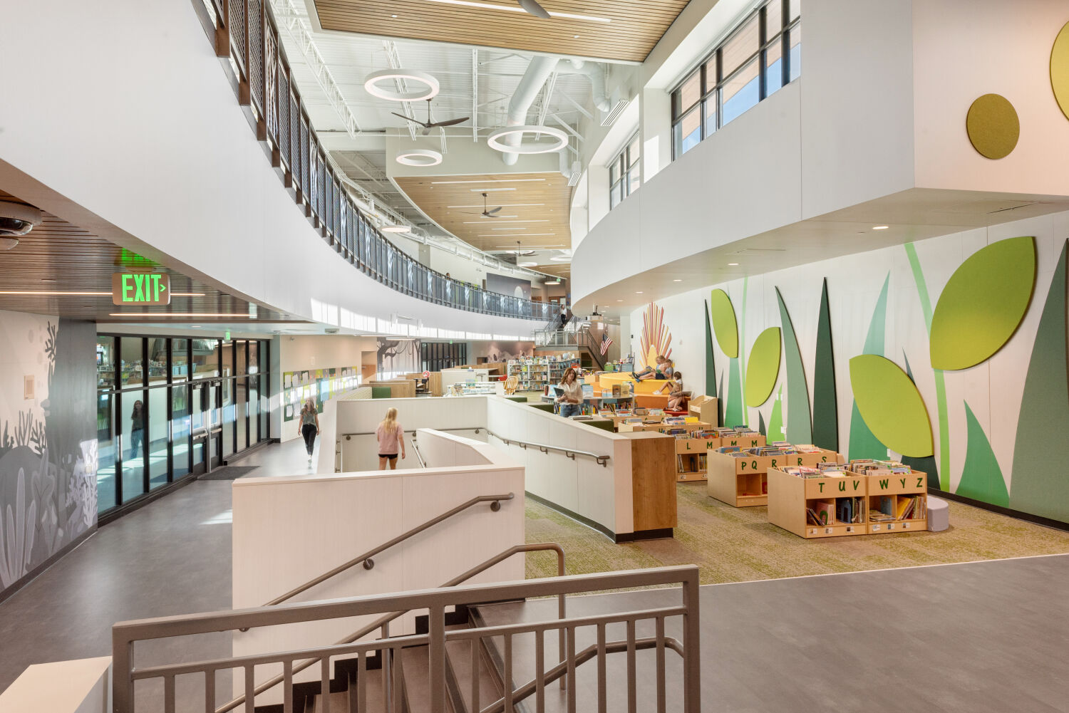 Two‑story media center with a curved mezzanine walkway, wood‑slat ceiling accents, circular pendant lights, large glass exterior wall, built‑in book displays, reading nooks, and a nature‑themed wall with oversized leaf graphics.