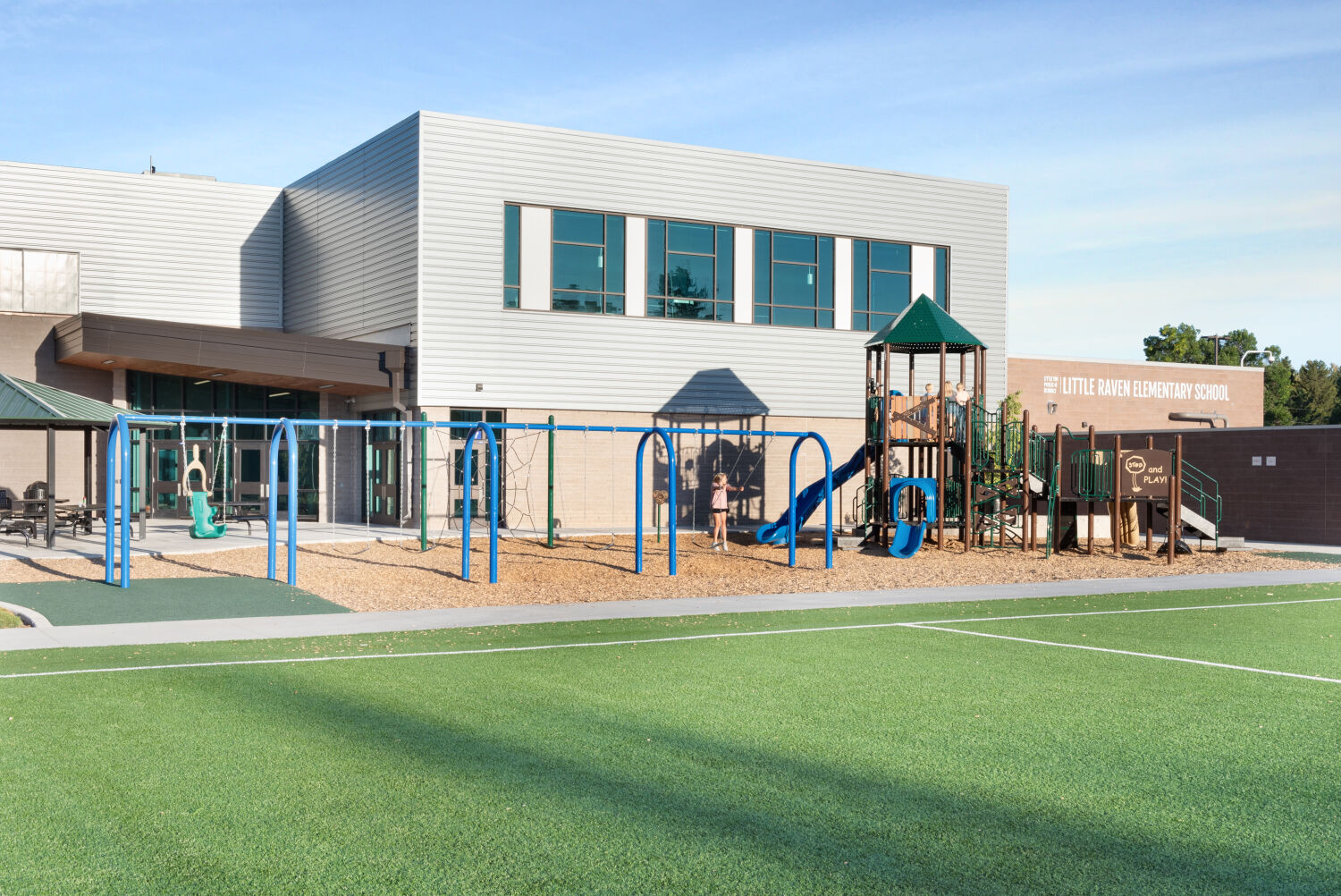 Exterior of Little Raven Elementary School with a two‑story building, adjacent playground with swings, climbing structure, blue slide, and wood‑mulch surfacing, next to a synthetic turf play field.