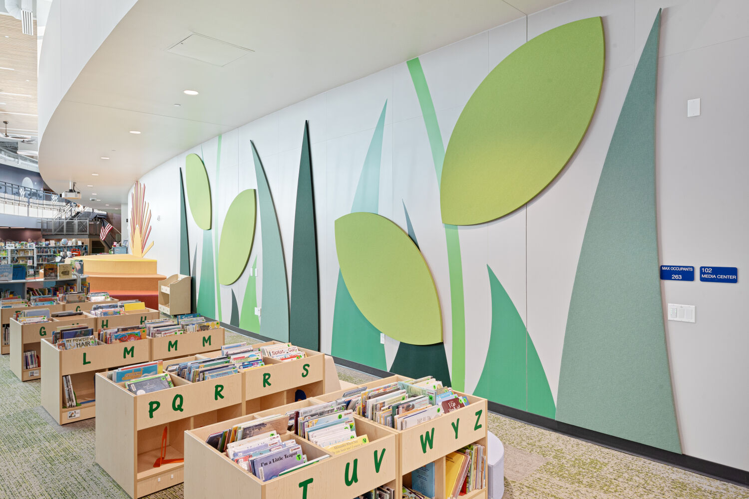 Media center with low book bins labeled by alphabet letters, green‑and‑gray carpet tile flooring, and a large leaf‑themed wall installation featuring oversized green leaf shapes and tall stem‑like forms along a curved interior wall.