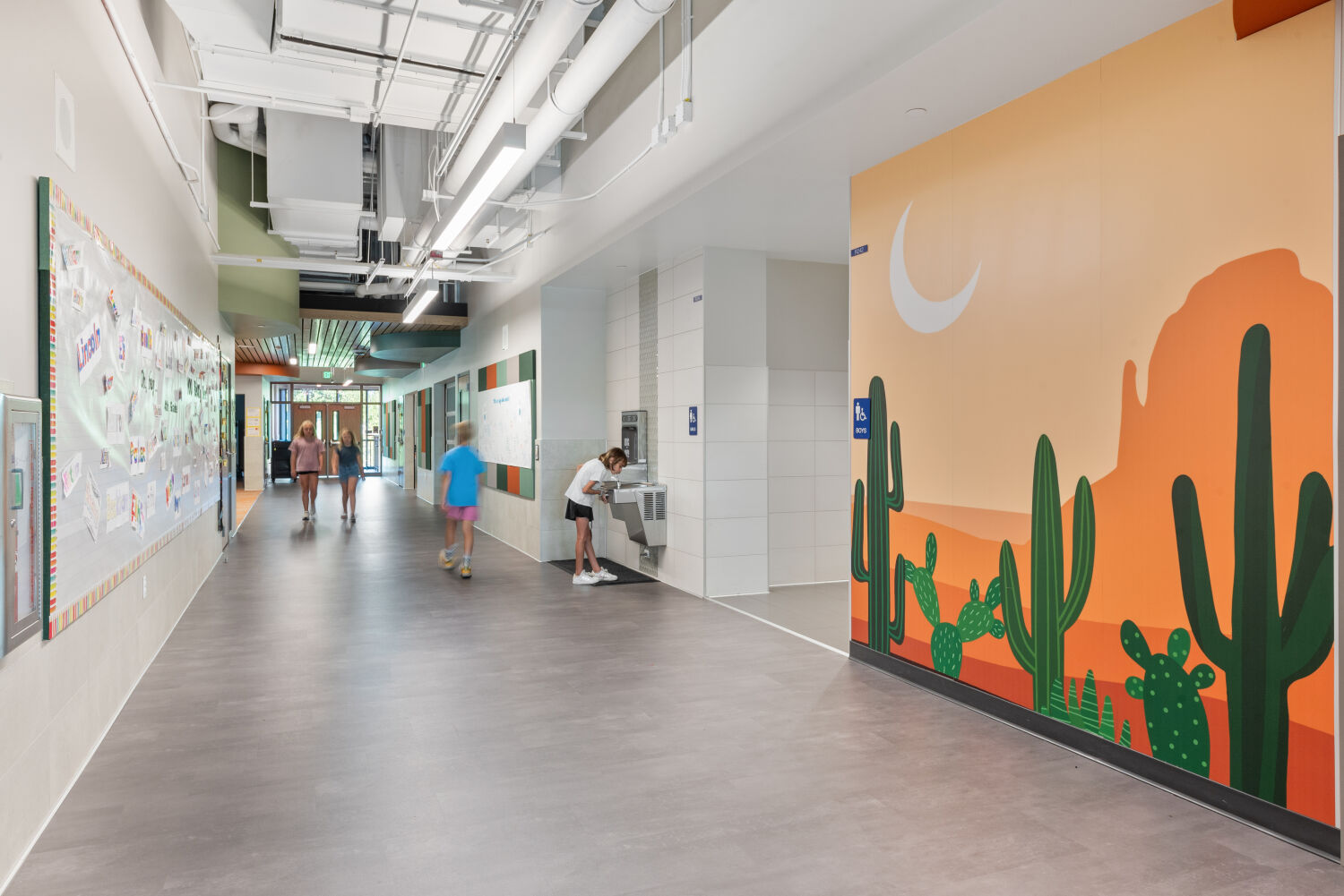 Interior corridor with a desert‑themed wall mural featuring illustrated cacti and mountains beneath a crescent moon, adjacent bulletin boards with student work, tiled drinking‑fountain alcove, and gray flooring.