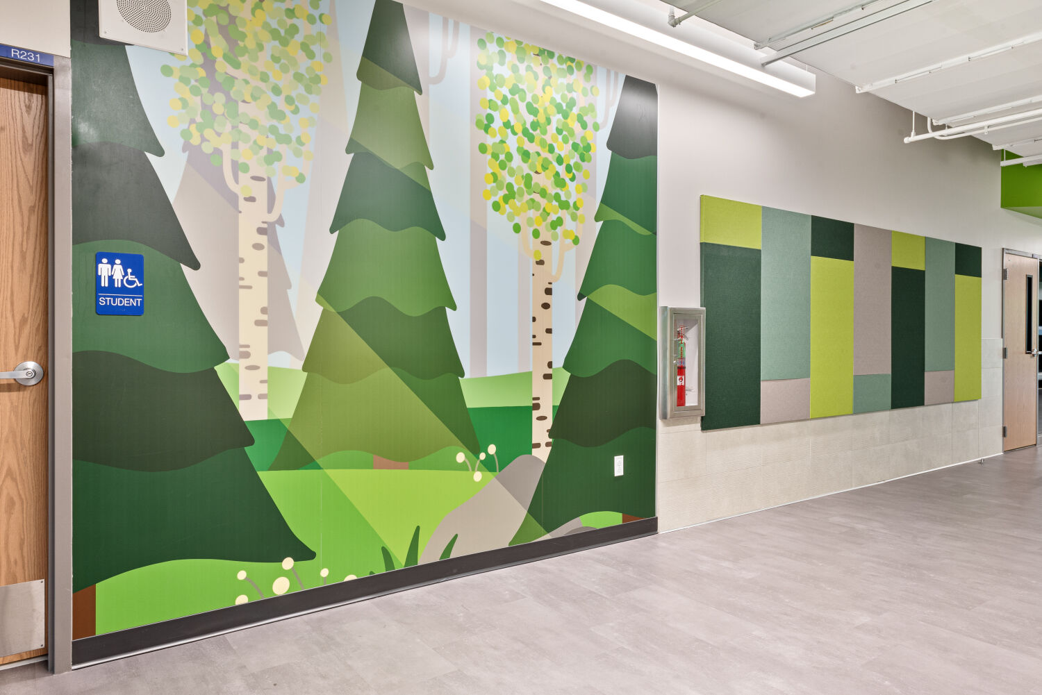 Interior corridor with a large forest‑themed wall mural featuring illustrated evergreen trees, birch trunks, and layered green foliage, adjacent to a wall of green and gray acoustic panels, and gray flooring.