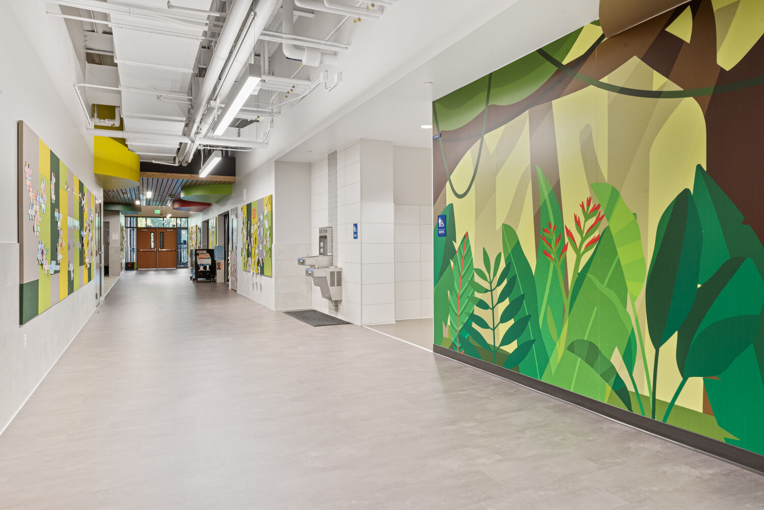 Interior corridor with a large jungle‑themed wall mural featuring oversized green foliage and tree shapes, adjacent bulletin boards with layered paper displays, and gray flooring leading toward double doors at the far end.