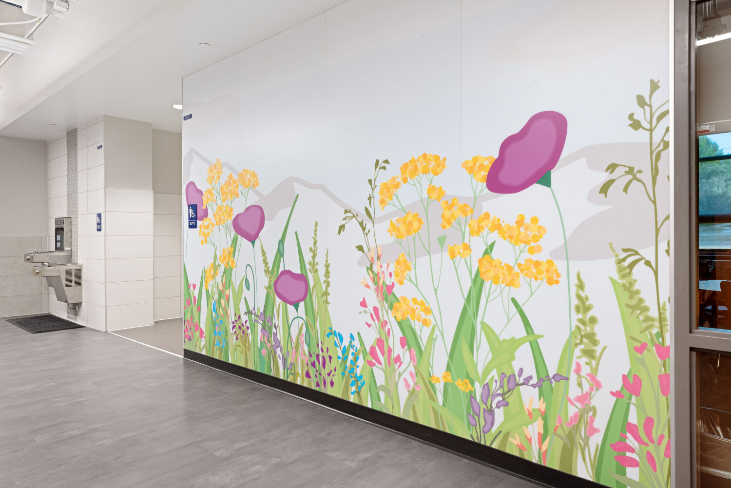 Interior corridor with a prairie‑themed wall mural featuring illustrated tall grasses, wildflowers, and soft mountain silhouettes in the background, adjacent to tiled wall surfaces, drinking fountains, and gray flooring.