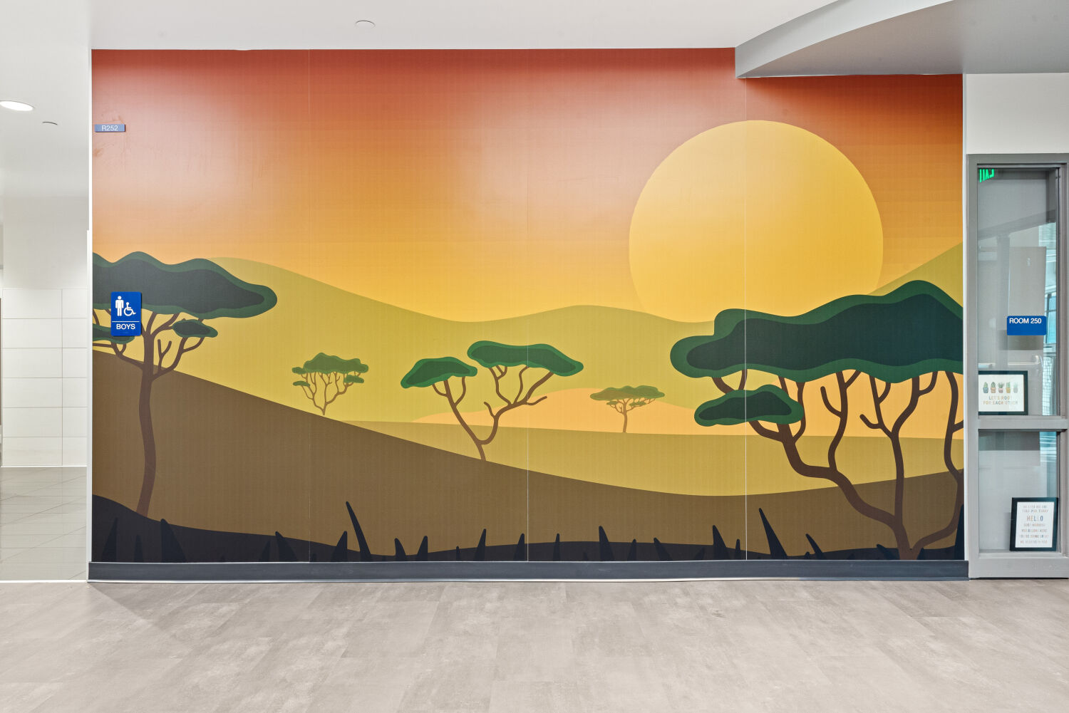 Interior corridor with a large savanna‑themed wall mural featuring silhouetted acacia trees, rolling hills, and a large setting sun in warm orange and yellow tones, adjacent to a glass‑framed doorway.