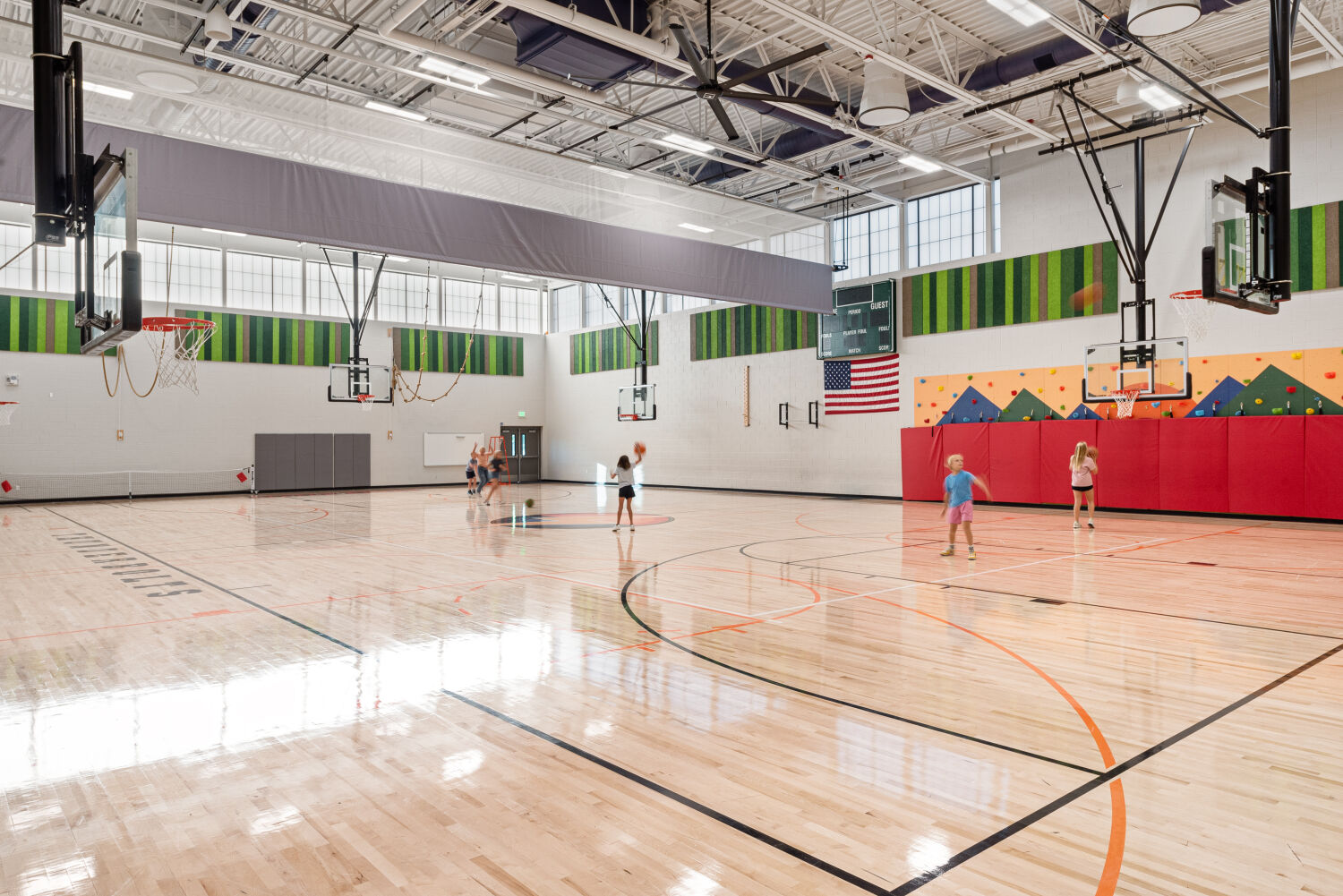 Gymnasium with a hardwood basketball court, multiple ceiling‑mounted hoops, wall padding in red and orange geometric patterns, striped acoustic wall panels in green tones, and high windows providing natural light.