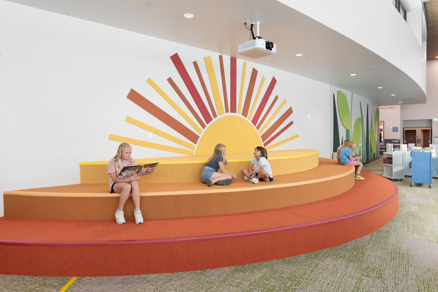 Tiered seating platform in the media center with curved orange and yellow steps forming a sunburst backdrop on the wall, surrounded by nature‑themed graphics and adjacent shelving and work areas.