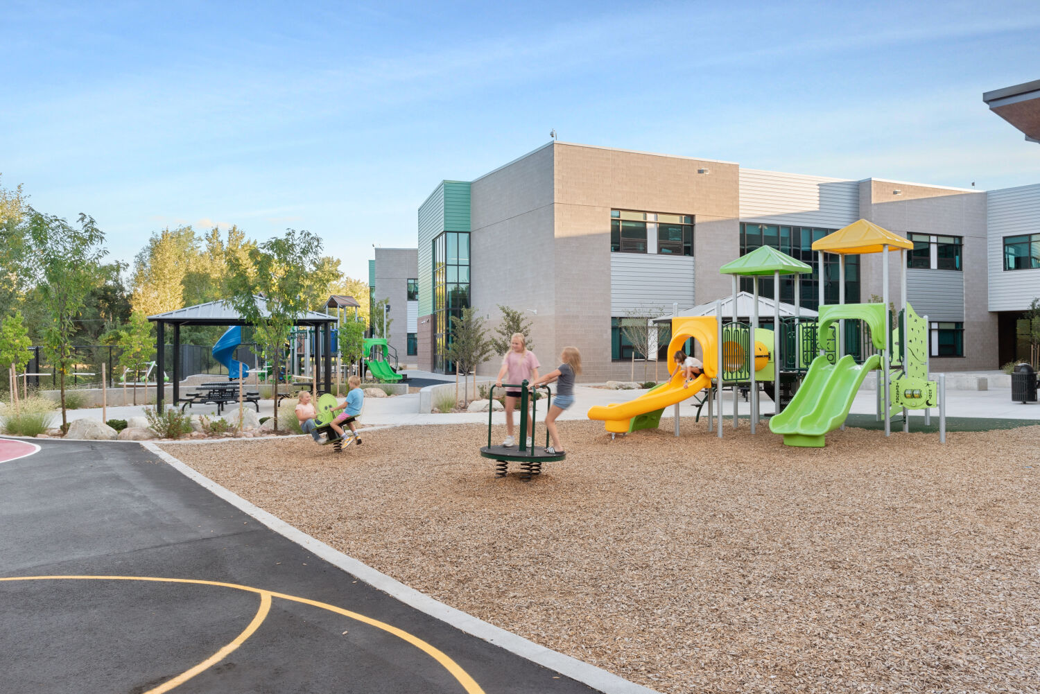 Outdoor play area with a paved game court featuring circular play markings, adjacent playground structures with slides and climbing elements, wood‑mulch surfacing, perimeter fencing, and surrounding trees and landscaping.