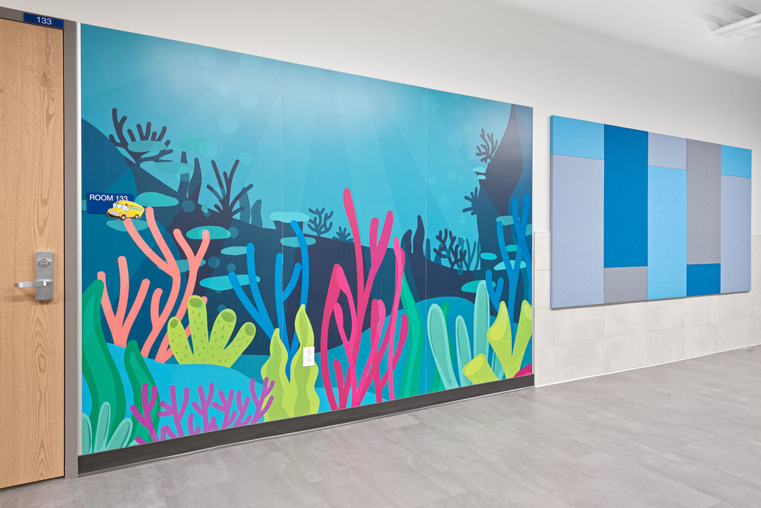 Interior corridor with a large under‑the‑sea themed wall mural featuring colorful coral, sea plants, and a small yellow submarine, adjacent to a wall of blue and gray acoustic panels and a wood‑framed classroom door.