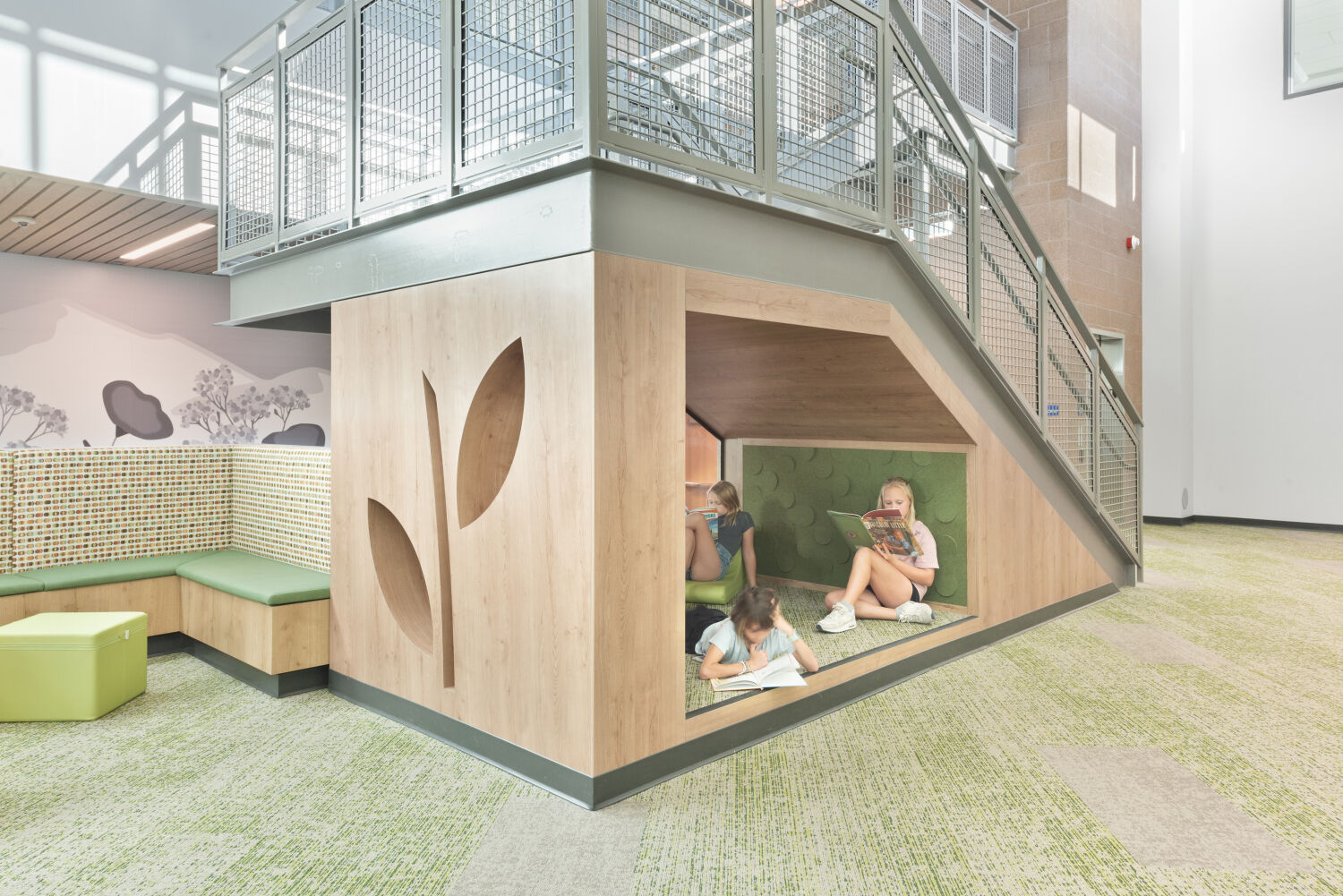 Under‑stair reading nook with wood paneling featuring carved leaf designs, green cushioned interior seating, adjacent built‑in bench seating with patterned upholstery, metal stair railing above, and carpet flooring in green and beige tones.